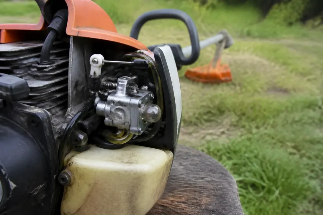 Plan rapproché d'une débroussailleuse thermique sur fond herbeux. Moteur et carburateur visibles.