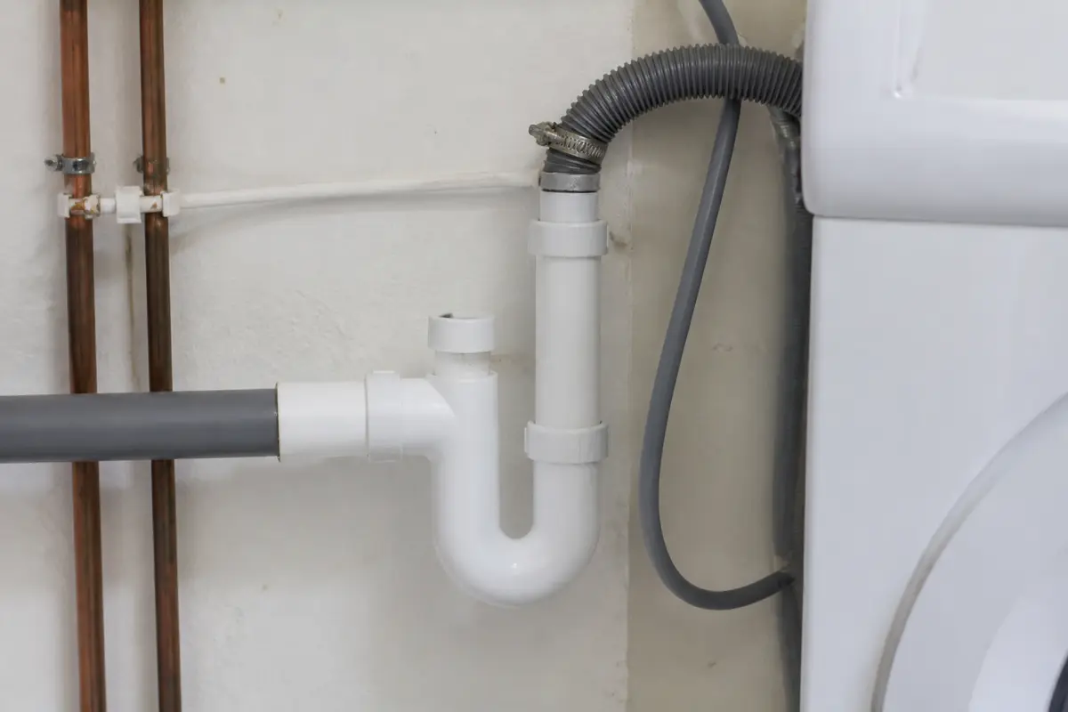 Tuyau de plomberie en PVC connecté à un mur et une machine, fixé par un collier métallique.