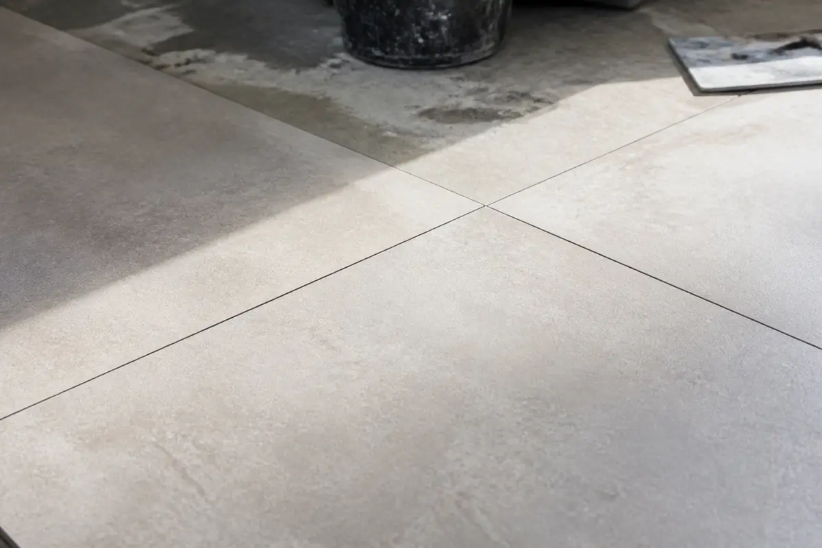 Carrelage gris clair, jointures fines, lumière naturelle, reflet ombre, sol en cours de pose, sceau au fond.