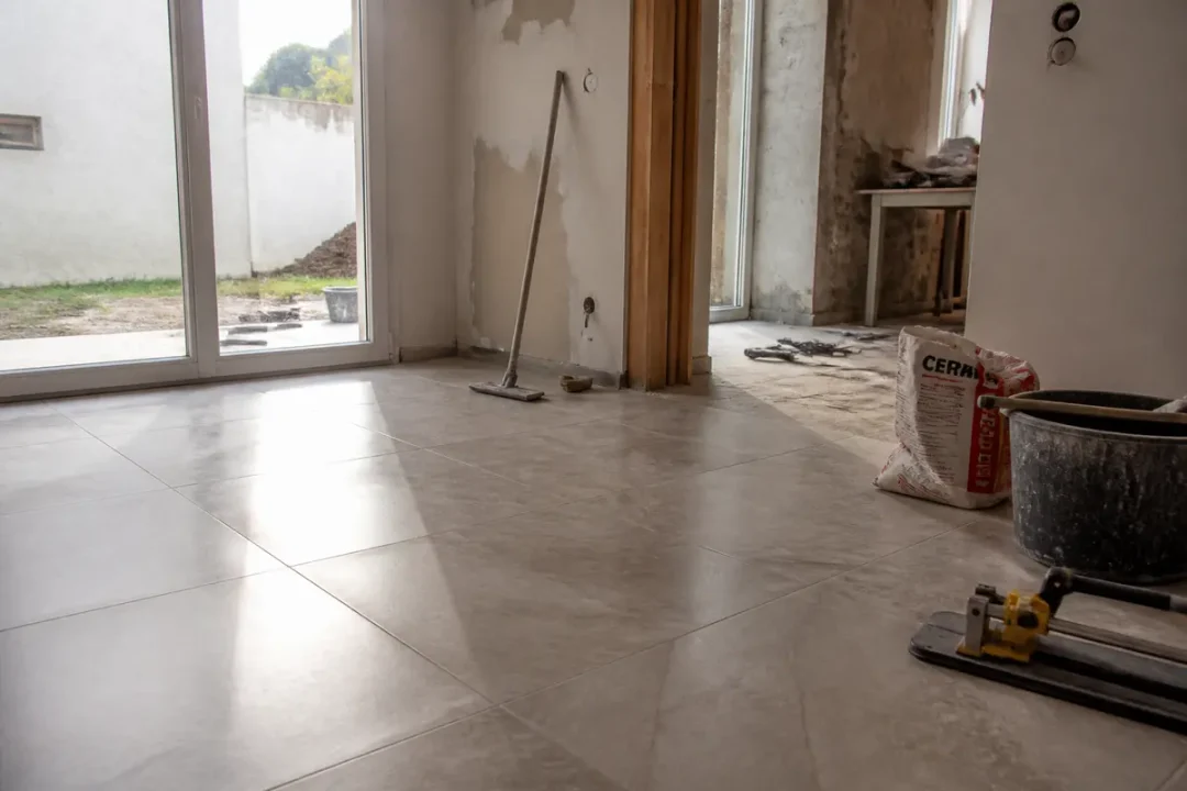 Intérieur en rénovation avec carrelage, outils, et lumière entrant par une baie vitrée. Sol refait, murs endommagés.