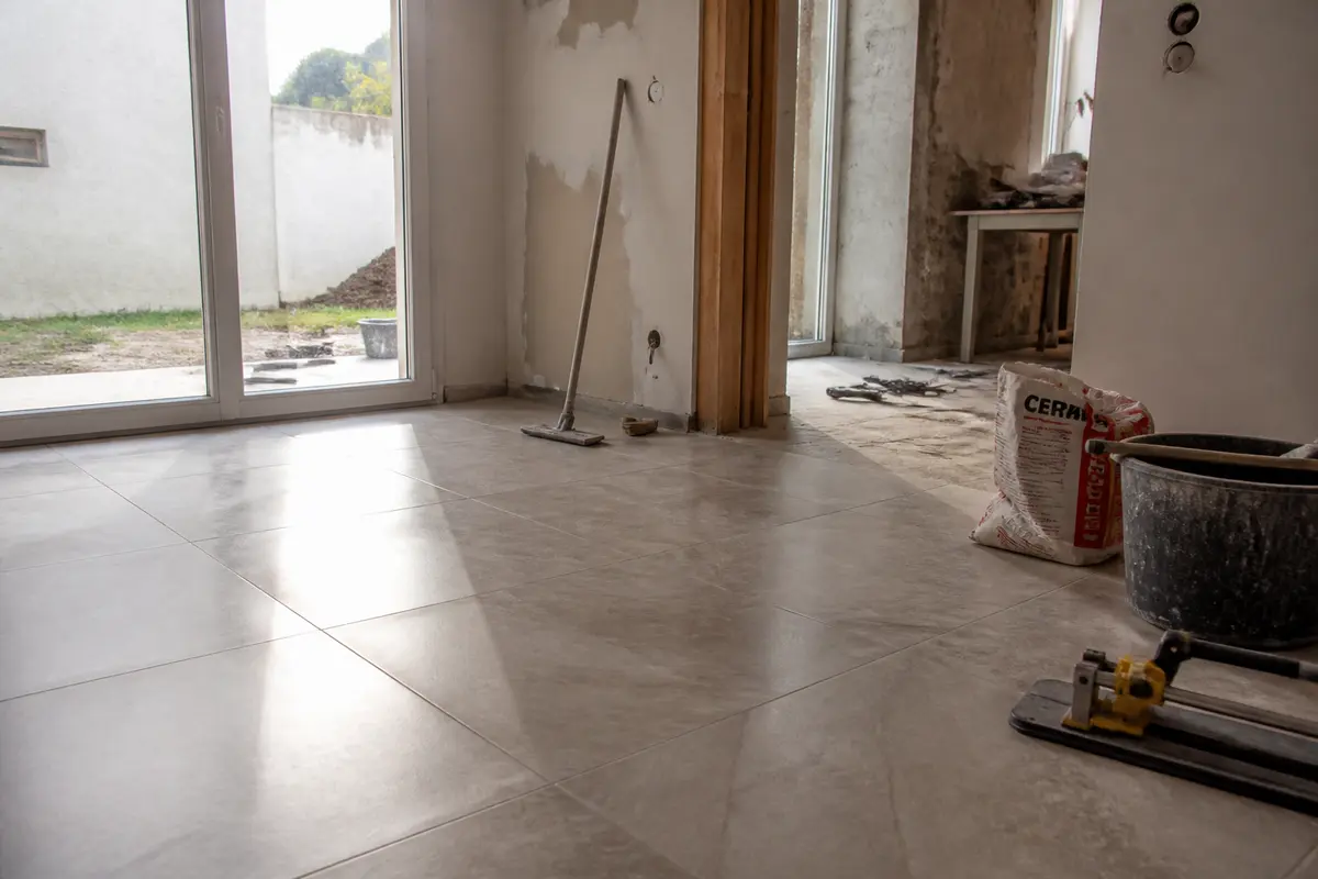 Intérieur en rénovation avec carrelage, outils, et lumière entrant par une baie vitrée. Sol refait, murs endommagés.