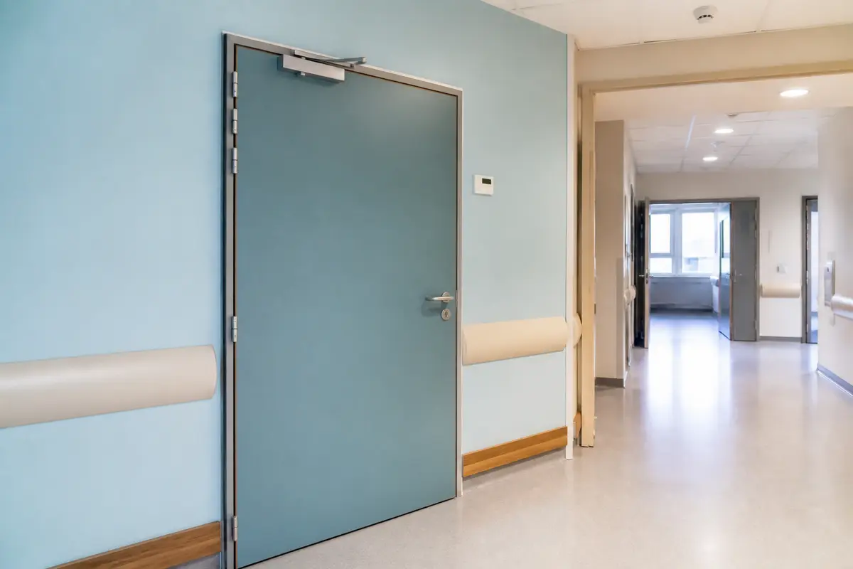 Couloir de milieu médical avec murs bleus, porte fermée, sol clair et lumière naturelle en arrière-plan.