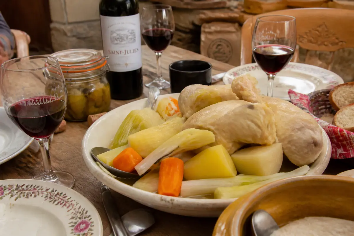 Plat de poule au pot avec légumes, verres de vin rouge, pain, et bouteille de vin sur table rustique.