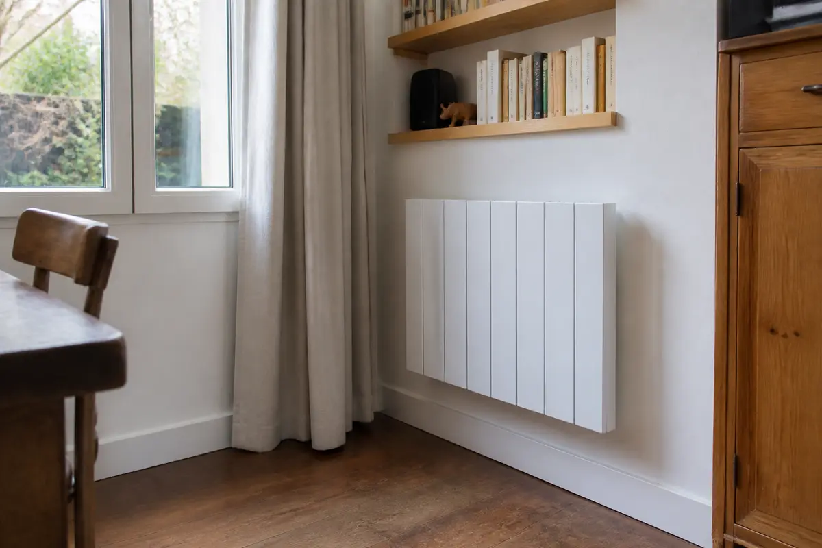 Radiateur blanc fixé à un mur blanc, près d'une fenêtre et d'une étagère avec des livres.