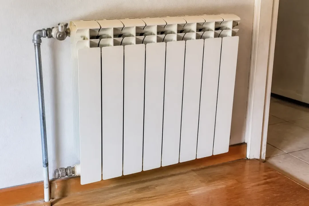 Radiateur blanc mural, installé sur plancher en bois, à côté d'une porte avec tuyaux de raccordement visibles.