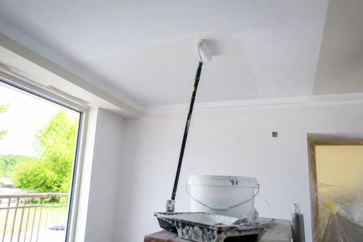 Un plafond est peint à rouleau dans une pièce claire avec seau et plateau. Fenêtre à gauche.