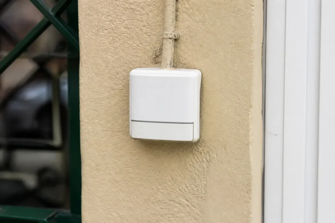 Boîtier blanc fixé sur un mur beige avec cadre de fenêtre vert à gauche.