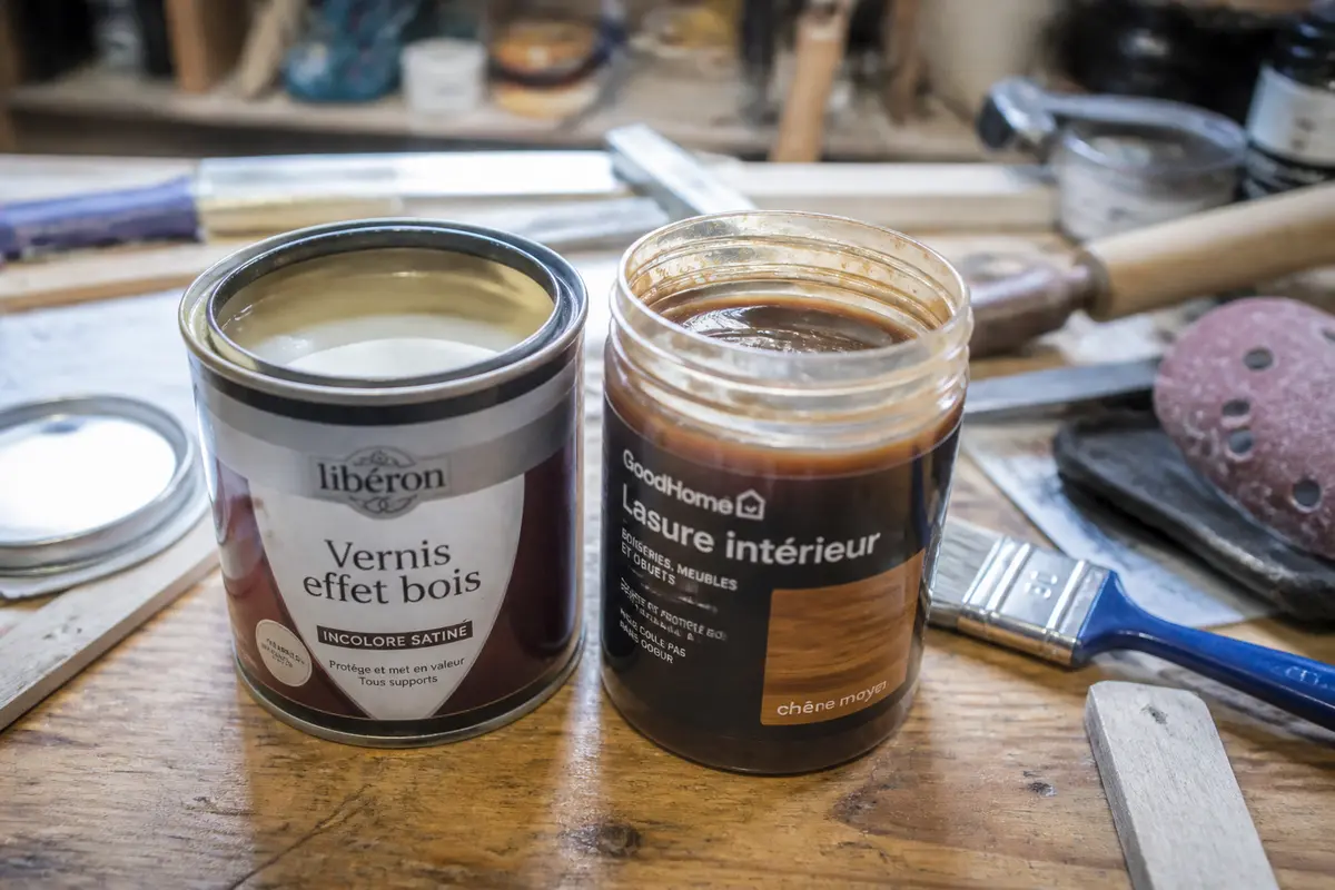 Deux pots de vernis et lasure en bois sur établi avec pinceau et outils à proximité.