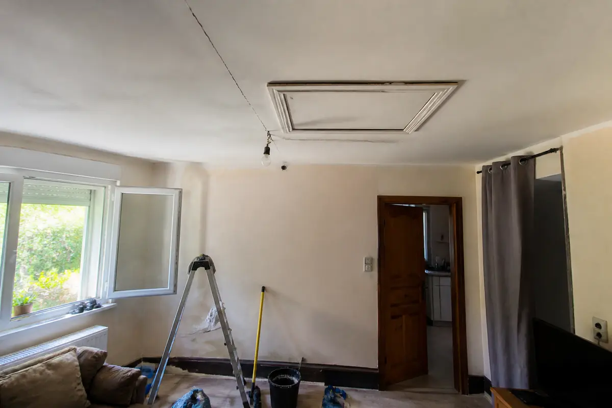 Salon en rénovation avec fenêtre ouverte, escabeau, outils et une pièce attenante visible à travers une porte.